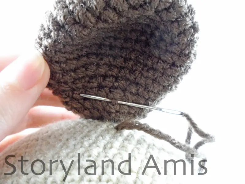 A close-up showing a hand lifting a crocheted dog ear on an amigurumi dog. A silver tapestry needle is inserted through a stitch on the underside of the ear.