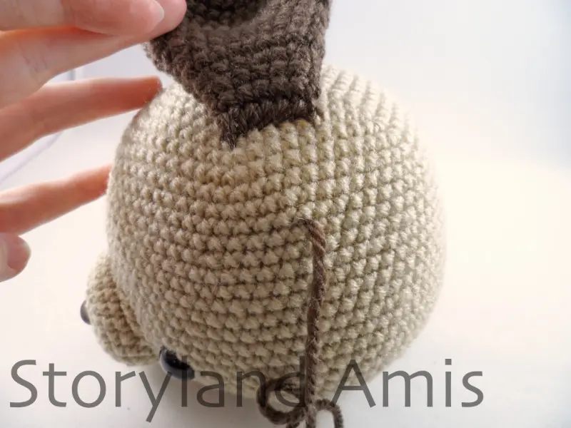 A close-up of the side of an amigurumi dog's head, with a brown yarn tail coming out of the side. A hand lifts the dog's ear up so that you can see underneath. 