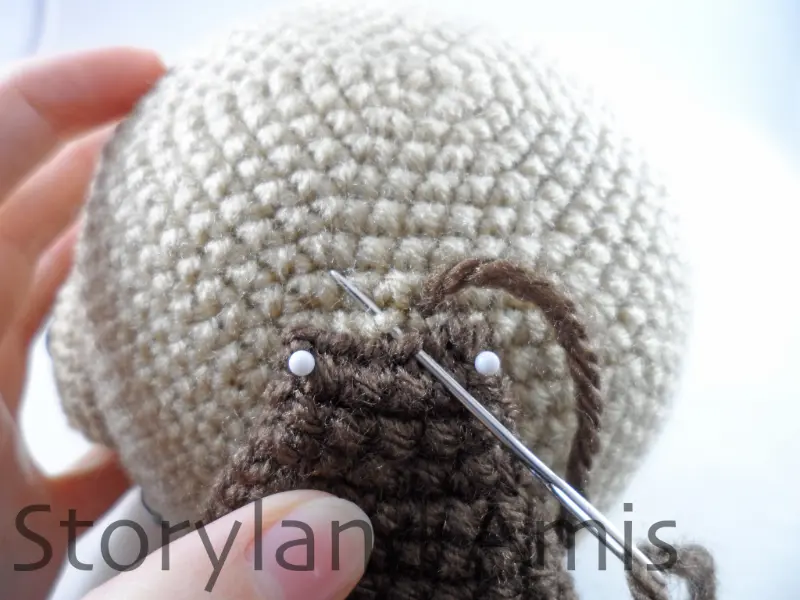 A close-up of a crocheted ear being sewn to an amigurumi dog's head using a tapestry needle.
