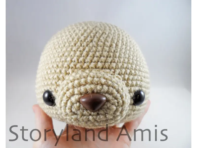 A hand holds up a partially finished amigurumi dog head. The dog has two plastic safety eyes and a crocheted muzzle sewn on with a brown plastic safety nose inserted in the center of it. 