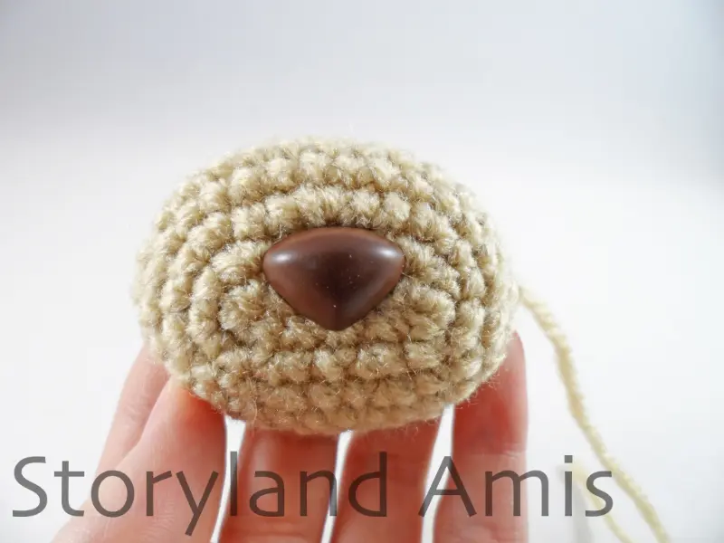 A hand holds up a tan crocheted muzzle with a brown plastic safety nose inserted in it for an amigurumi dog.