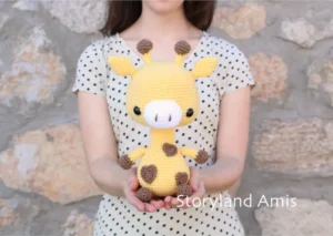A girl wearing a tan shirt with black polka dots stands in front of a rock wall holding a crocheted yellow and brown amigurumi giraffe.