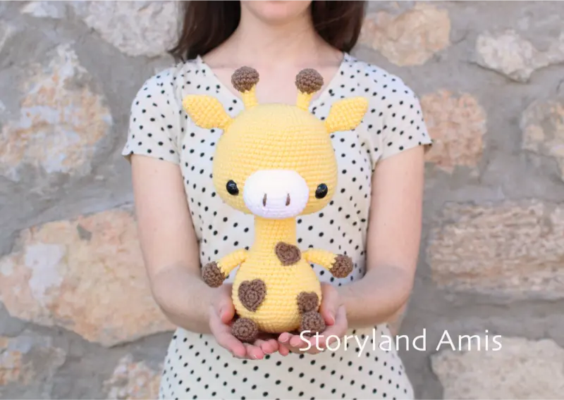 A girl wearing a tan shirt with black polka dots stands in front of a rock wall holding a crocheted yellow and brown amigurumi giraffe.