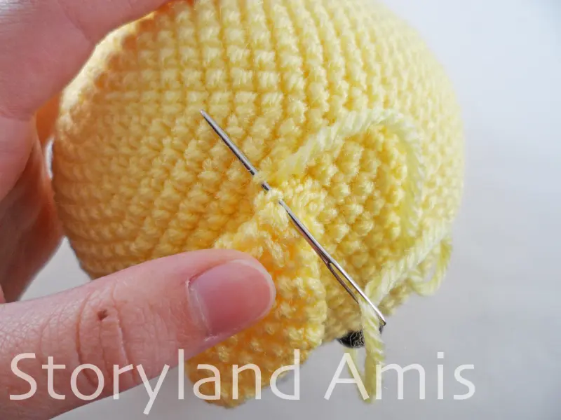 A hand uses a silver tapestry needle to sew a crocheted ear to the head of an amigurumi giraffe toy.
