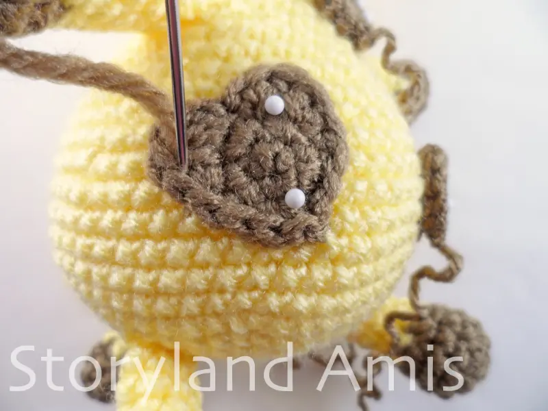 A close-up of a silver tapestry needle being used to sew a brown crocheted heart applique to the body of an amigurumi giraffe.