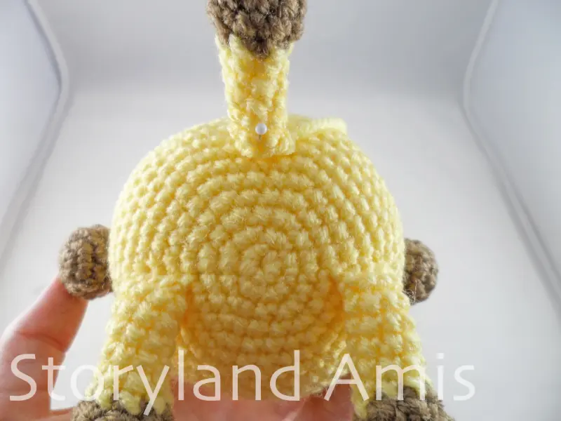 A close-up of the bottom of a crocheted amigurumi giraffe showing the tail pinned to the back with straight pins.
