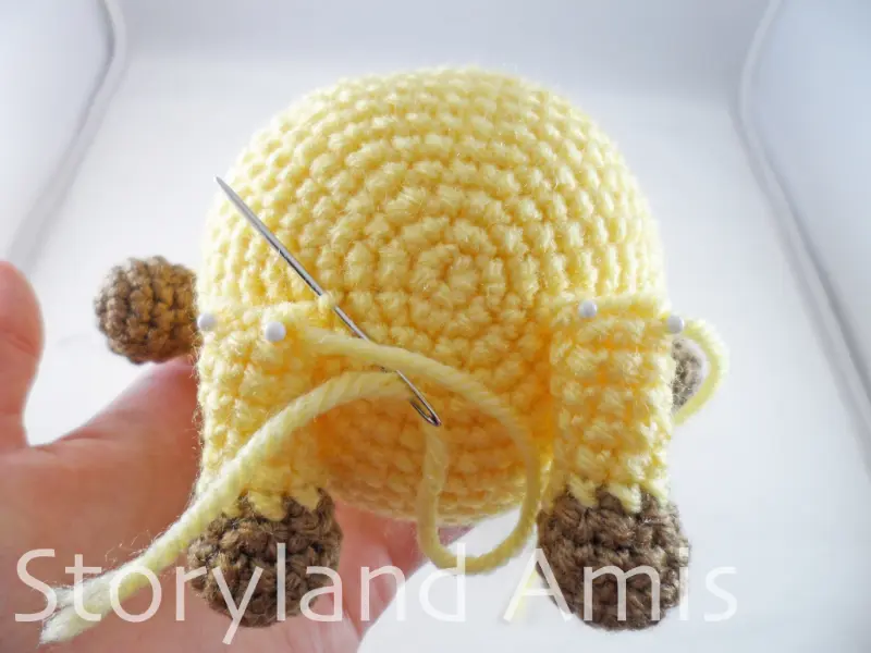 A close-up of the bottom of an amigurumi giraffe showing the legs pinned to the body with straight pins. A tapestry needle is inserted through the stitches of the left leg and body.