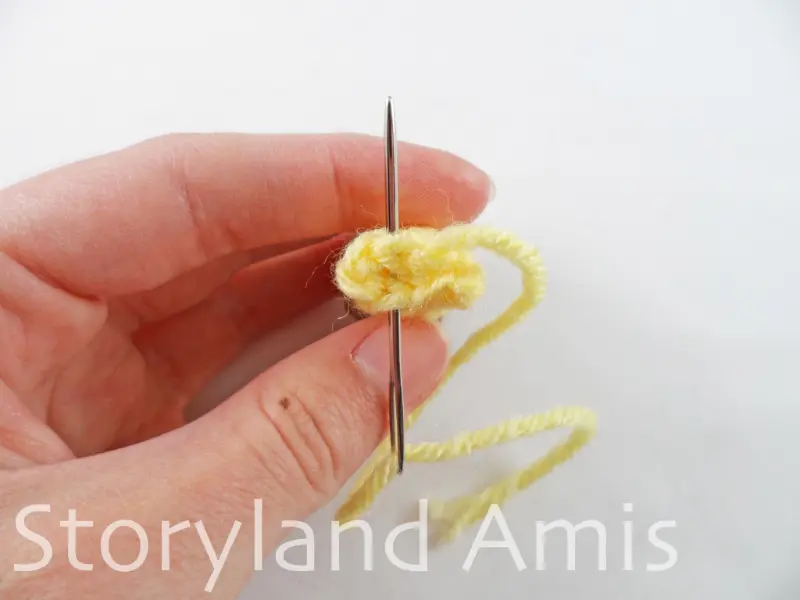 A hand holds a small crocheted yellow giraffe ear. A silver tapestry needle is inserted in the bottom stitches of the ear.