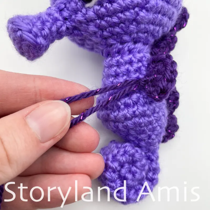 A hand holds two dark purple yarn tails coming from the body of a purple amigurumi seahorse toy lying on a white background.