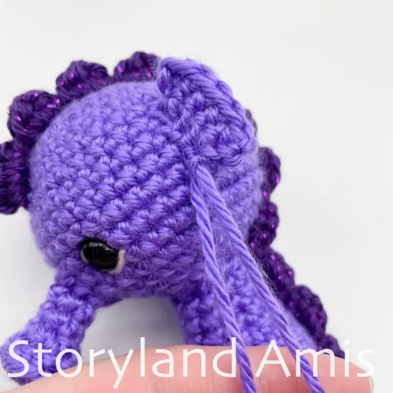 A close-up of an amigurumi seahorse's head with two purple yarn tails coming out of the head.