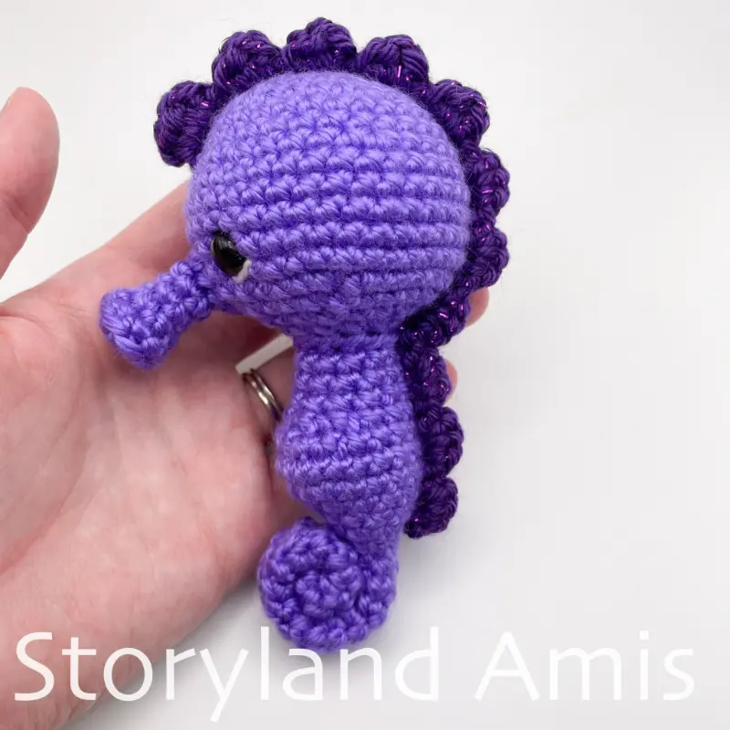 A hand holds a partially finished amigurumi seahorse against a white background.