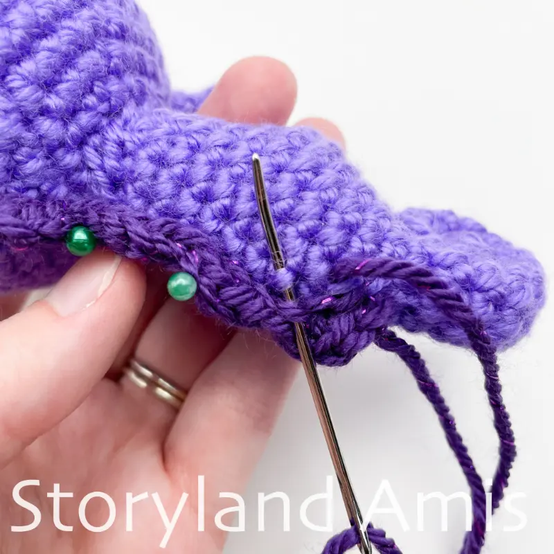 A close-up of a hand using a tapestry needle to sew a dark purple fringe to the back of a purple amigurumi seahorse.