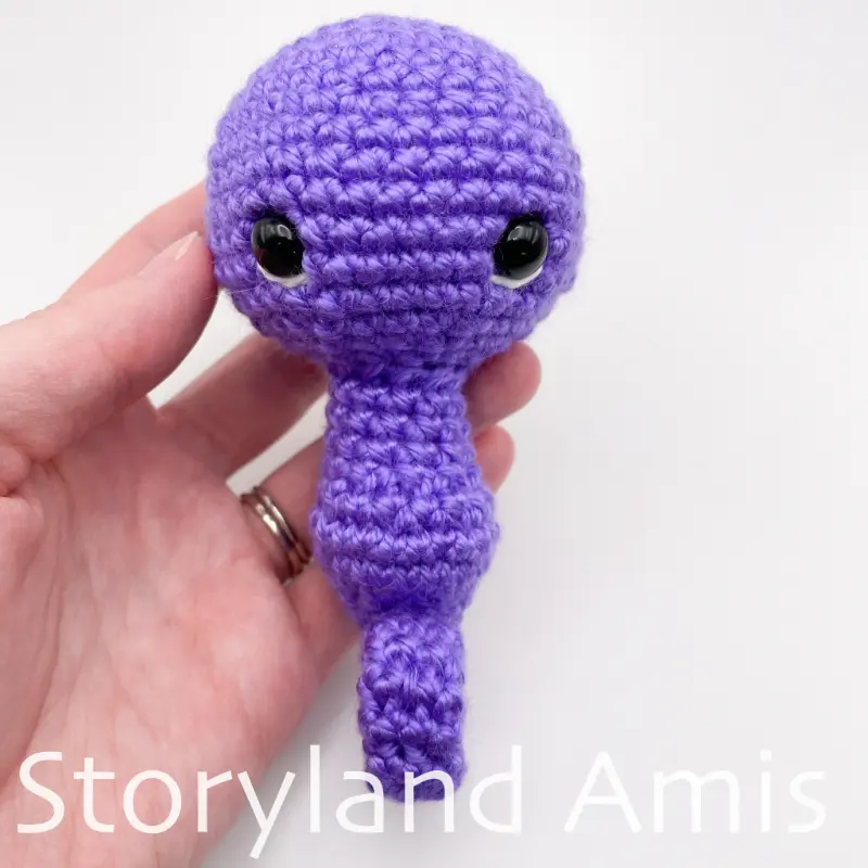 A hand holds a partially completed purple crocheted seahorse against a white background.