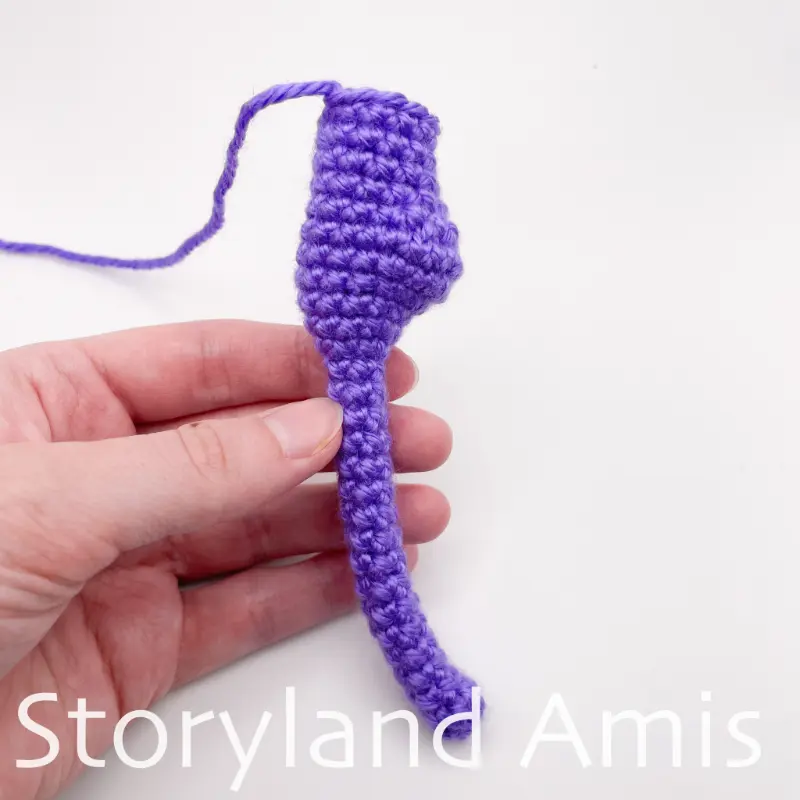 A hand holds a purple crocheted seahorse body against a white background.
