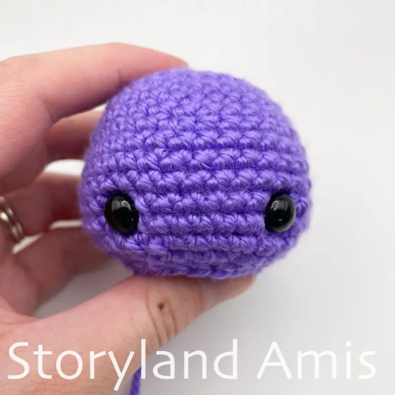 A hand holds a crocheted purple amigurumi seahorse head.