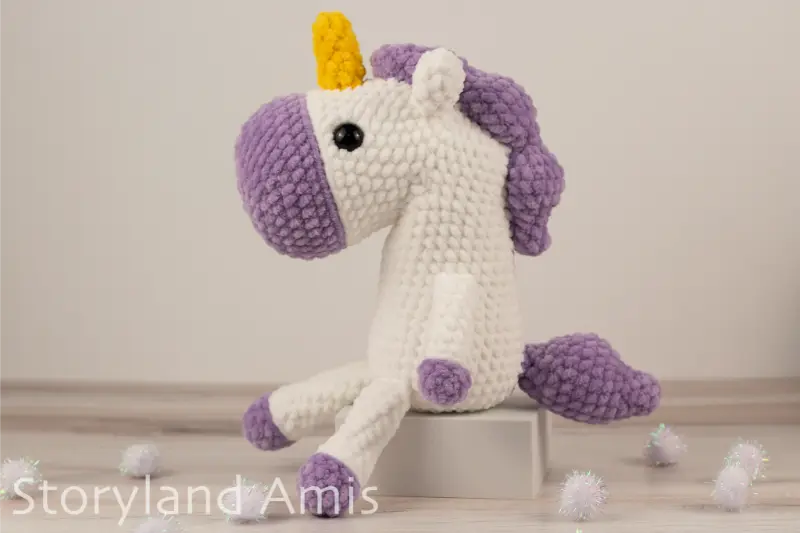 A crocheted white and purple unicorn sits on a small white box sitting on a wooden table. White pom poms are scattered around the table.