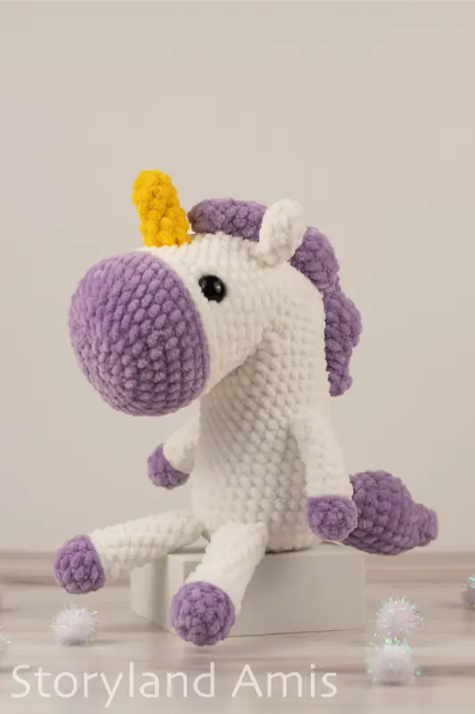 A crocheted white and purple unicorn sits on a small white box sitting on a wooden table. White pom poms are scattered around the table.