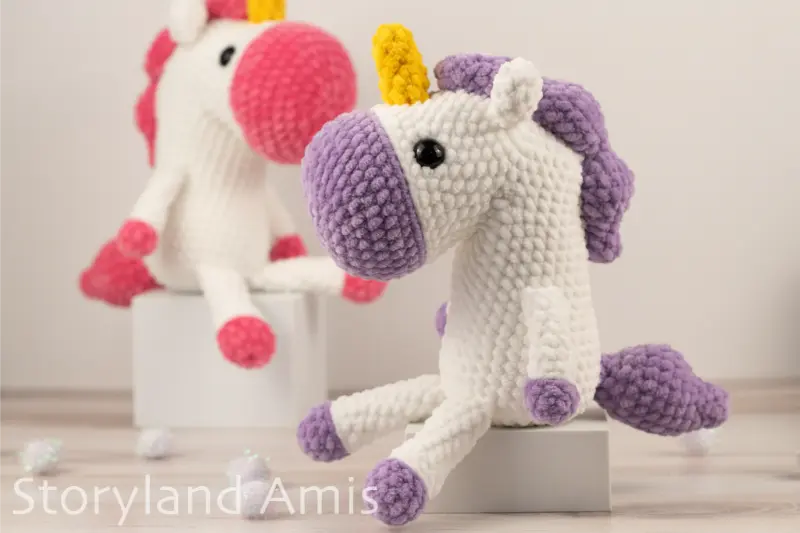 Two crocheted unicorns sit on small white boxes placed on a wooden table. White pom poms are scattered around the table.