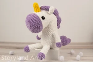 A crocheted white and purple unicorn sits on a small white box sitting on a wooden table. White pom poms are scattered around the table.