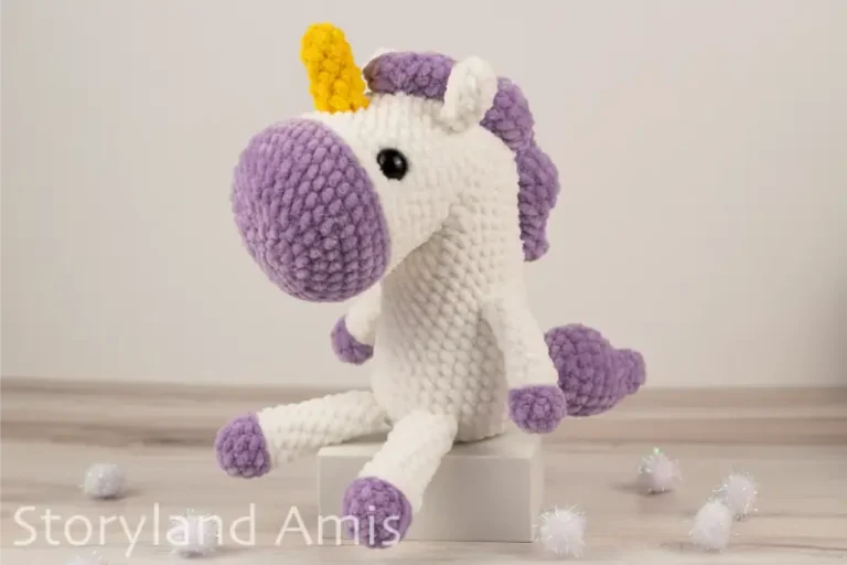 A crocheted white and purple unicorn sits on a small white box sitting on a wooden table. White pom poms are scattered around the table.