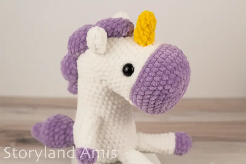 A crocheted white and purple unicorn sits on a white box placed on a wooden table.