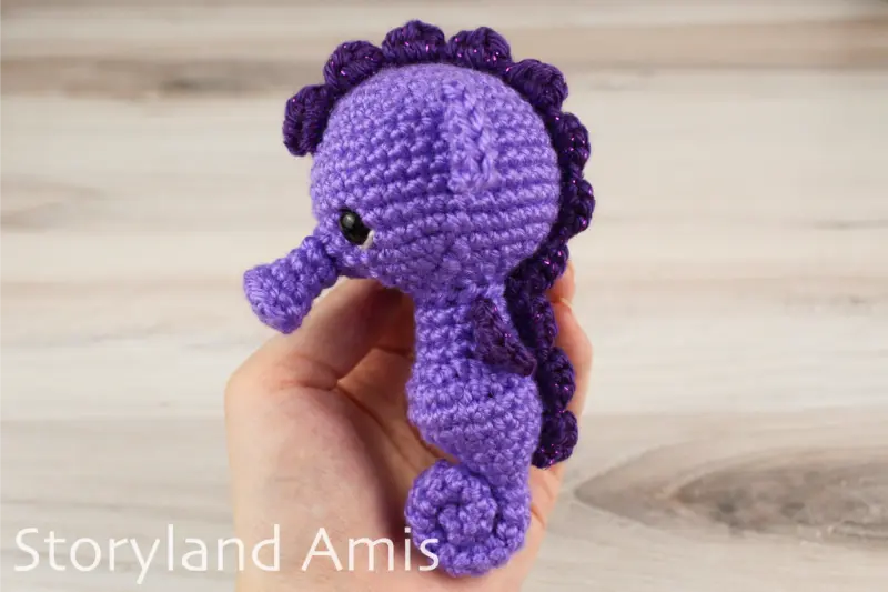 A hand holds a purple amigurumi seahorse against a wooden backdrop.