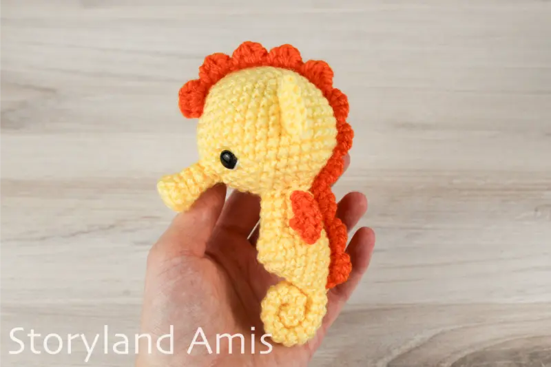 A hand holds a crocheted yellow and orange seahorse against a wooden background.