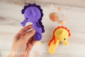 A hand holds a purple crocheted seahorse against a wooden background where another yellow and orange seahorse lies. There are seashells scattered over the table.