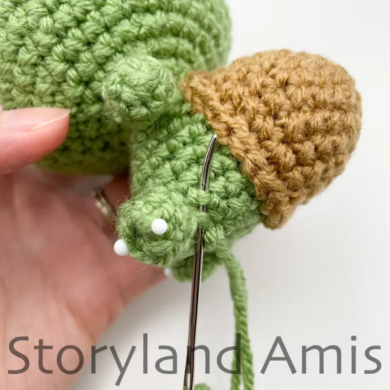 A close-up of a tapestry needle being used to sew a small leg to the body of an amigurumi tortoise.