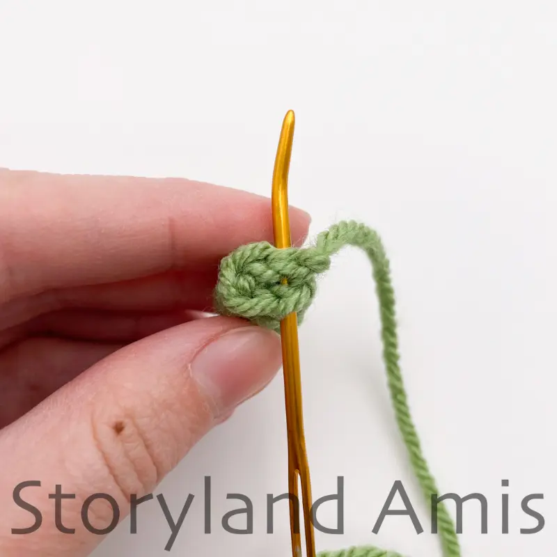 A hand holds a small, green crocheted arm. A tapestry needle is inserted through the stitches on the edge of the arm.