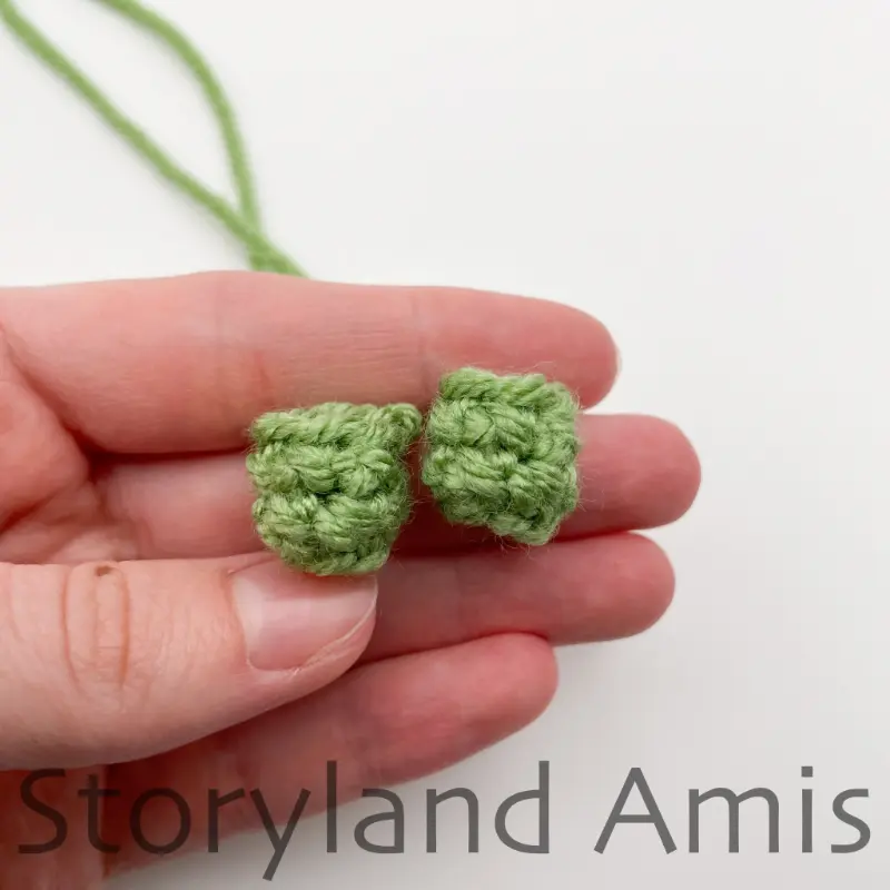 A hand holds two small, green crocheted legs for an amigurumi tortoise.