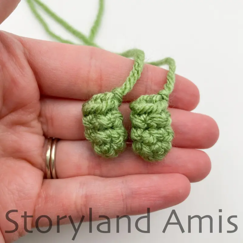 A hand holds two small, green crocheted arms for an amigurumi tortoise.