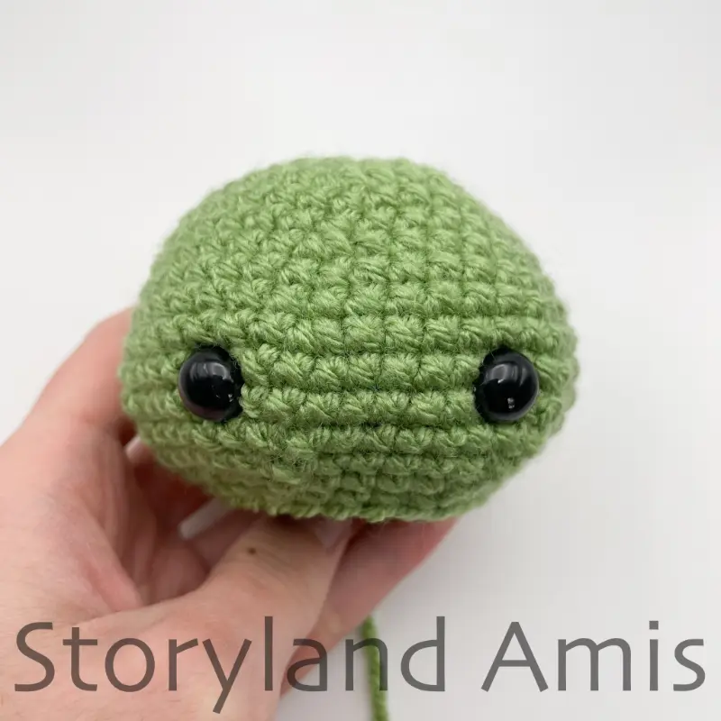 A hand holds a small, green crocheted tortoise head.