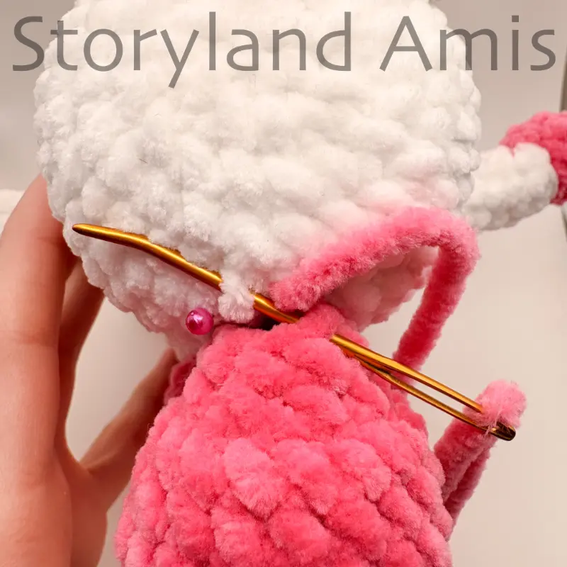 A close-up shows a tapestry needle being used to sew a pink crocheted tail to the back of an amigurumi unicorn's body.