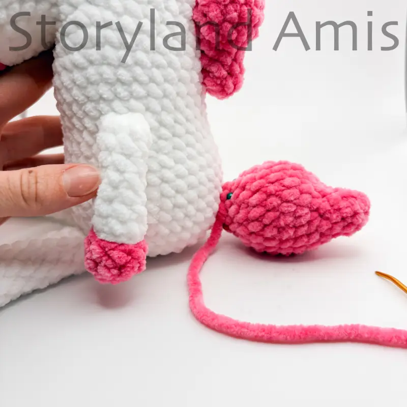 A close-up of a crocheted unicorn sitting on a white table with a pink tail pinned to its back with straight pins.