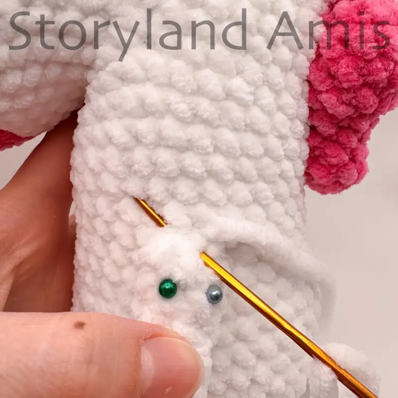 A close-up showing a tapestry needle being used to sew a crocheted arm to an amigurumi unicorn's body. The arm is pinned to the body with straight pins.