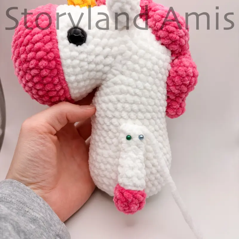 A hand holds a partially completed amigurumi unicorn with an arm pinned to it's side with straight pins.