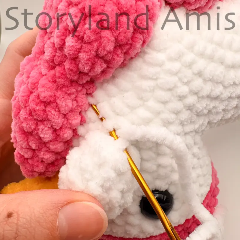 A close-up of a hand using a tapestry needle and yarn to sew a crocheted ear to an amigurumi unicorn's head.