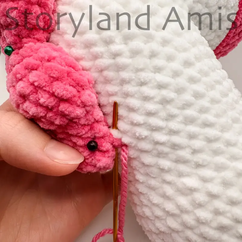A close-up of a hand using a tapestry needle and yarn to sew a pink mane to the body of an amigurumi unicorn toy.