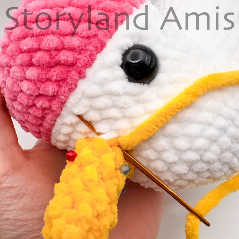A close-up of a gold tapestry needle being used to sew a crocheted yellow horn to an amigurumi unicorn's head.