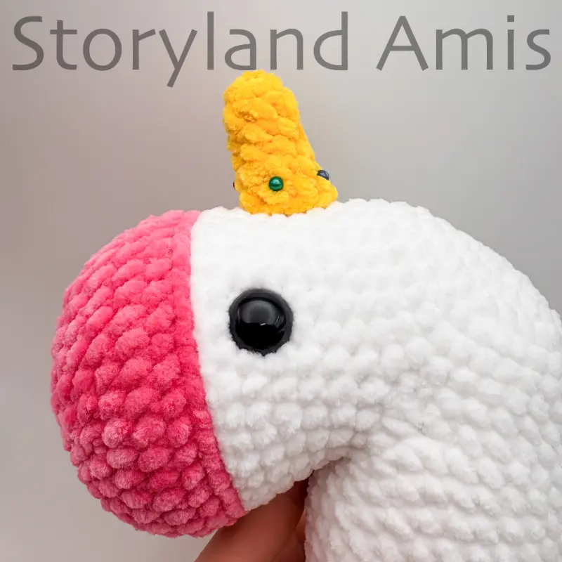 The side of an amigurumi unicorn's head with a crocheted yellow horn pinned between to the top of the head between the eyes.