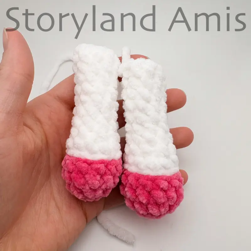 A hand holds two pink and white crocheted legs for an amigurumi unicorn.