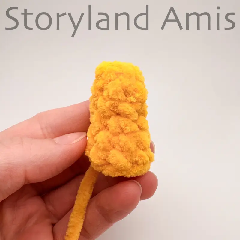 A hand holds up a small, yellow crocheted horn for an amigurumi unicorn toy.