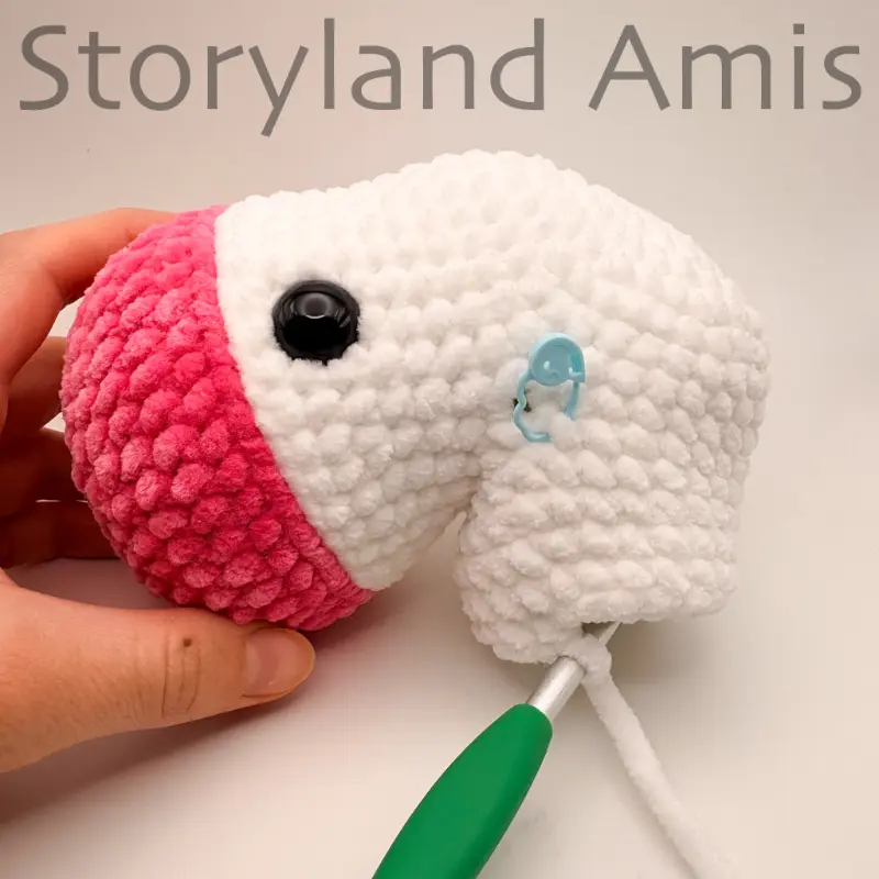 A hand steadies a partially crocheted amigurumi unicorn. A green crochet hook is inserted in a stitch of the body.