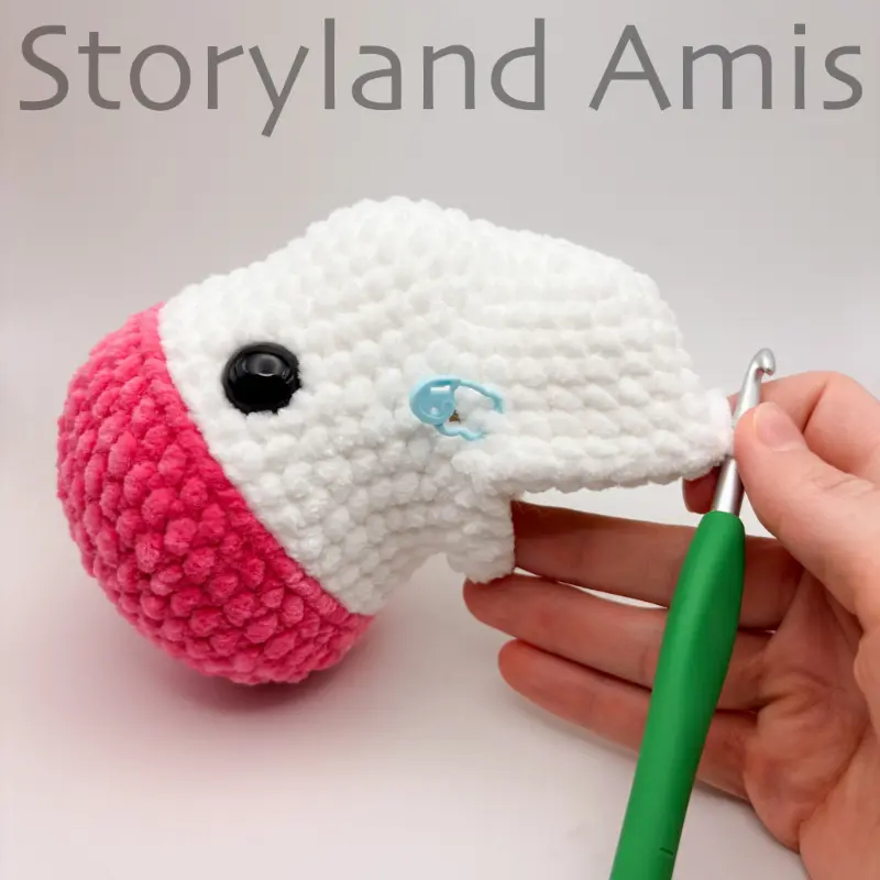 A hand steadies a partially crocheted amigurumi unicorn. A green crochet hook is inserted in a stitch of the body.