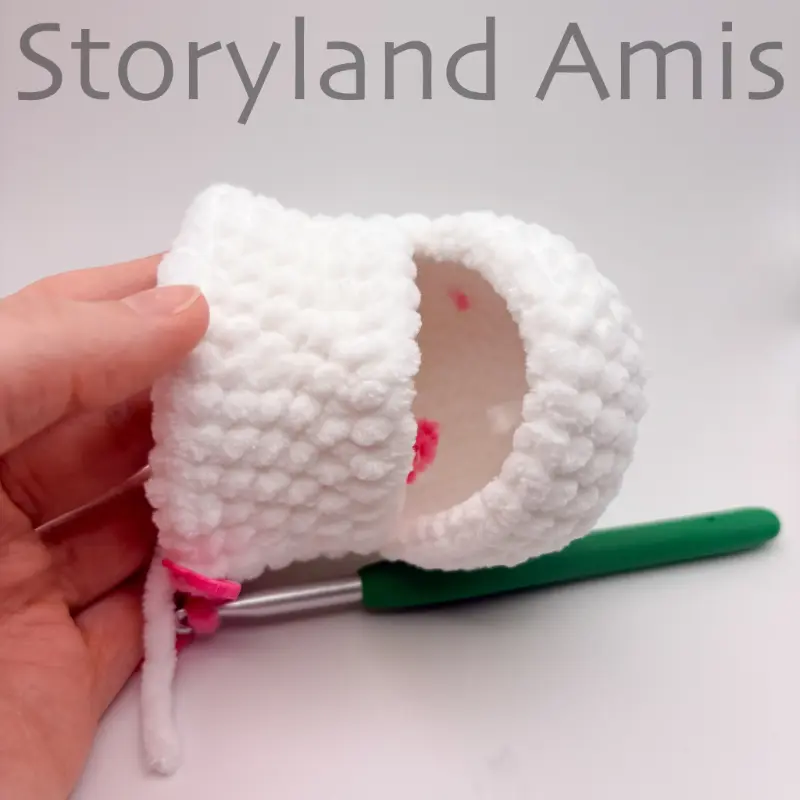 A partially crocheted amigurumi unicorn head is held by a hand. A green crochet hook is inserted in a stitch of the head.