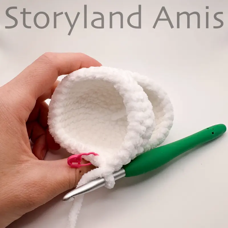 A partially crocheted unicorn head is held by a hand. A green crochet hook is inserted in a stitch of the head.