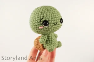 A hand holds a crocheted green and brown amigurumi tortoise against a white background.