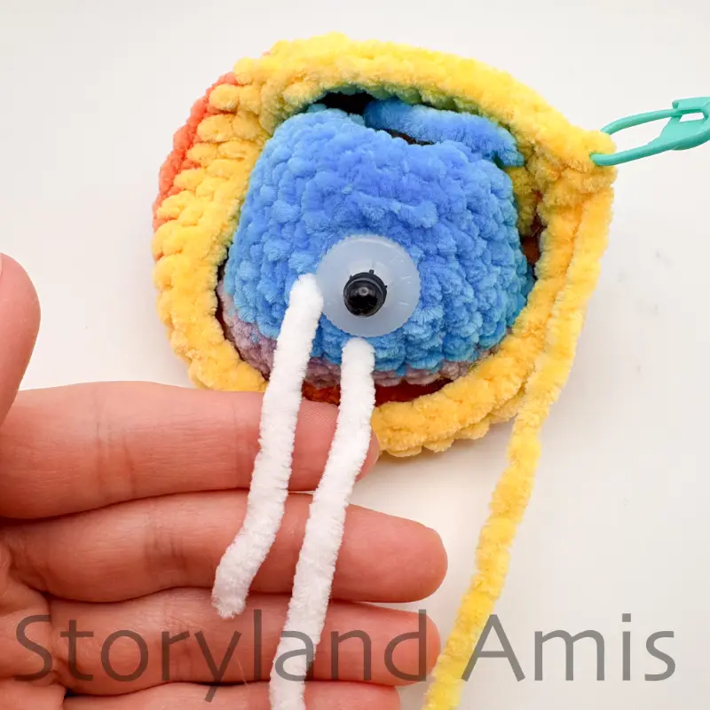 The inside of a partially finished crocheted fish lies on a white background. Two white yarn tails extend from the inside of the fish, draped over a hand.