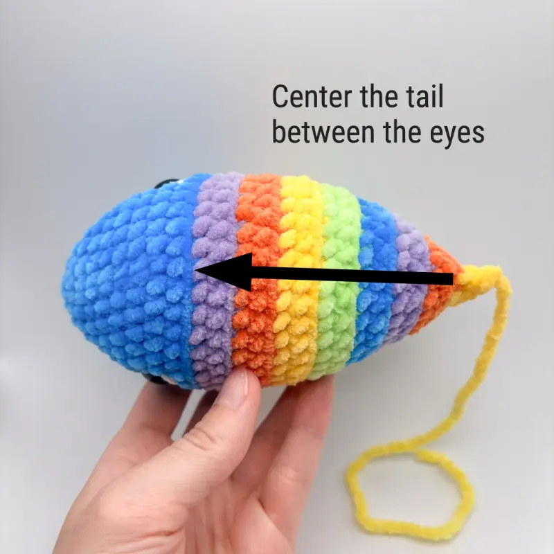 A hand holds an amigurumi fish, shown from the top. Text on the image reads, "Center the tail between the eyes". A black arrow overlays the back of the fish pointing in a straight line from the back of the tail to the front of the head.
